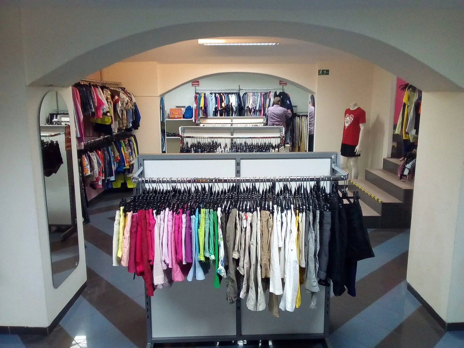 A clothing shop seen through an archway, with dense racks of garments on hangers and a mannequin on the right