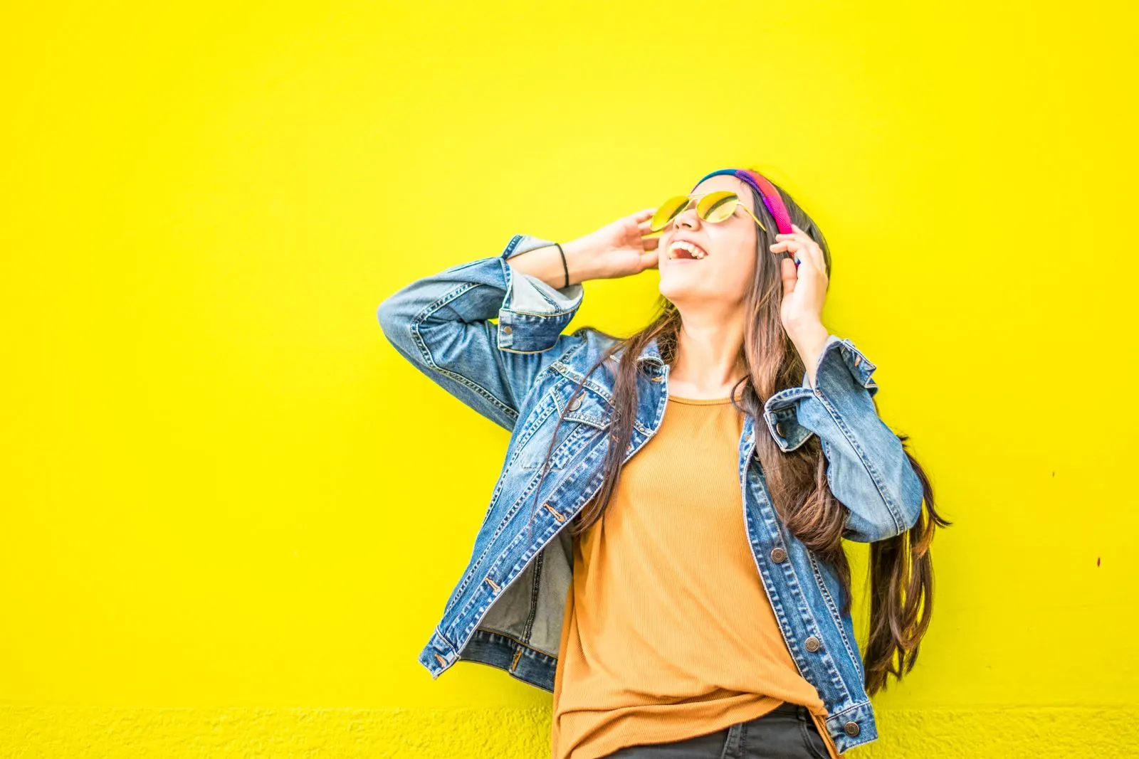 A woman in a denim jacket and warm orange top against a yellow wall