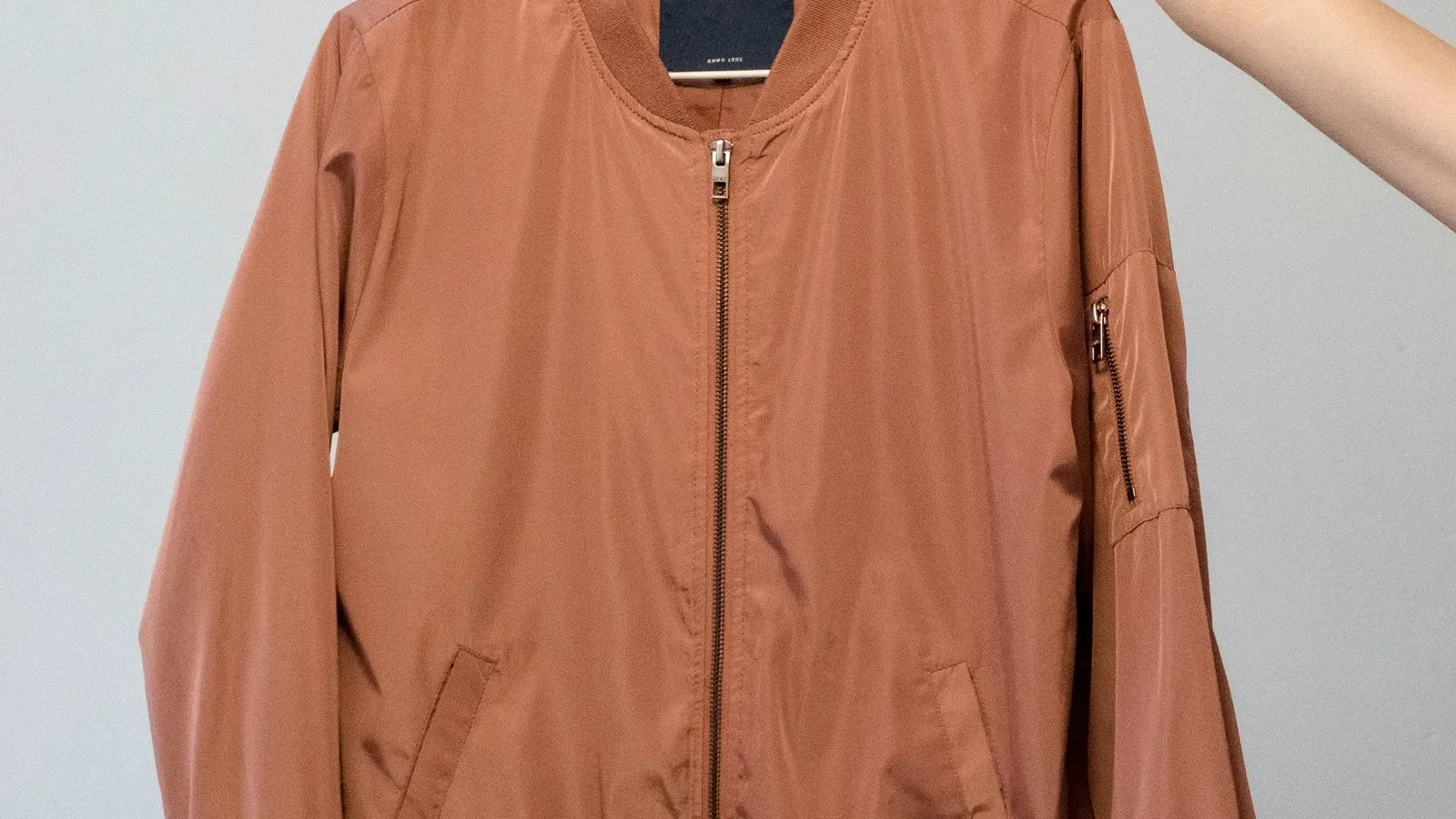 A light brown bomber jacket held on a hanger against a plain studio background