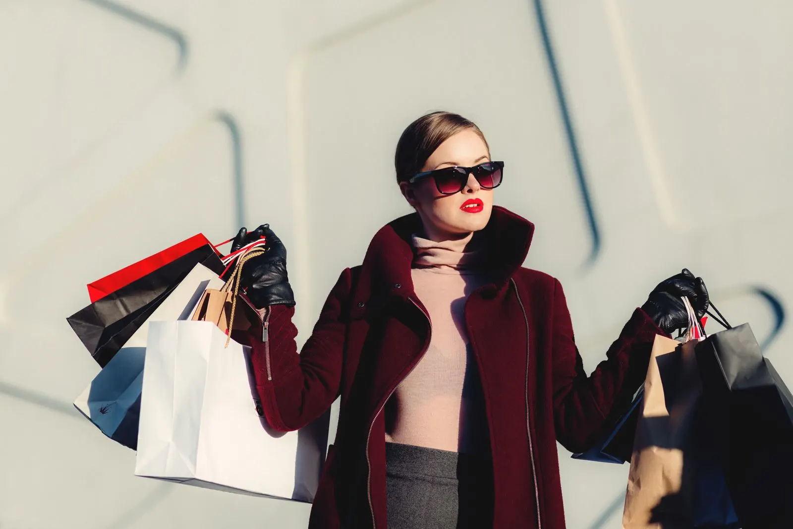 A woman carrying shopping bags in a deep red coat and black sunglasses